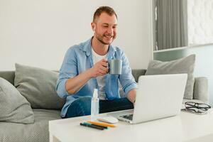 knap glimlachen Mens zittend Aan sofa drinken thee Bij huis Bij tafel werken online Aan laptop foto