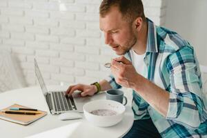 knap glimlachen Mens in overhemd zittend in keuken Bij huis Bij tafel werken online Aan laptop foto