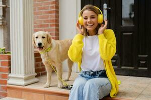 vrouw in geel trui wandelen Bij haar huis met een hond luisteren naar muziek- in hoofdtelefoons foto