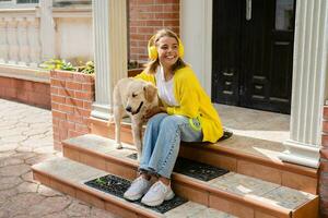 vrouw in geel trui wandelen Bij haar huis met een hond luisteren naar muziek- in hoofdtelefoons foto