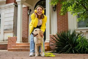 vrouw in geel trui wandelen Bij haar huis met een hond luisteren naar muziek- in hoofdtelefoons foto