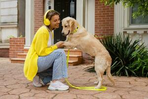 vrouw in geel trui wandelen Bij haar huis met een hond luisteren naar muziek- in hoofdtelefoons foto