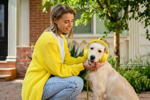 vrouw in geel trui wandelen Bij haar huis met een hond luisteren naar muziek- in hoofdtelefoons foto