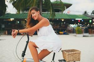 jong aantrekkelijk glimlachen vrouw in wit jurk rijden Aan tropisch strand Aan fiets foto