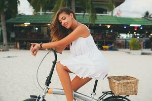 jong aantrekkelijk glimlachen vrouw in wit jurk rijden Aan tropisch strand Aan fiets foto