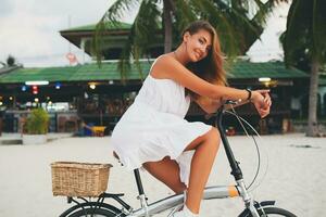 jong aantrekkelijk glimlachen vrouw in wit jurk rijden Aan tropisch strand Aan fiets foto