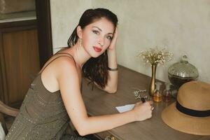 jong elegant mooi vrouw zittend Bij tafel in toevlucht hotel kamer, schrijven een brief, Holding pen foto