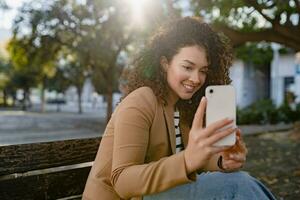 mooi gekruld glimlachen vrouw wandelen in stad straat in elegant jasje, gebruik makend van smartphone foto