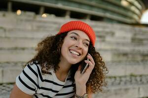 mooi gekruld glimlachen vrouw zittend in stad straat in gestreept t-shirt en gebreid rood hoed, gebruik makend van smartphone foto