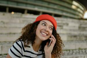 mooi gekruld glimlachen vrouw zittend in stad straat in gestreept t-shirt en gebreid rood hoed, gebruik makend van smartphone foto