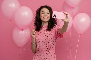 mooi jong vrouw poseren geïsoleerd Aan roze studio achtergrond met roze lucht baloons en smartphone foto