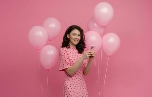 mooi jong vrouw poseren geïsoleerd Aan roze studio achtergrond met roze lucht baloons en smartphone foto