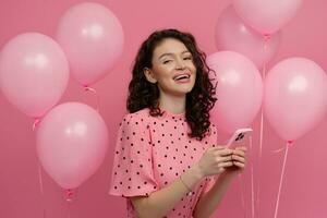 mooi jong vrouw poseren geïsoleerd Aan roze studio achtergrond met roze lucht baloons en smartphone foto