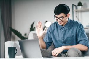 jonge aziatische man die smartphone gebruikt voor online videoconferentiegesprek zwaaiende hand hallo gebaar maken op de bank in de woonkamer 's nachts. foto