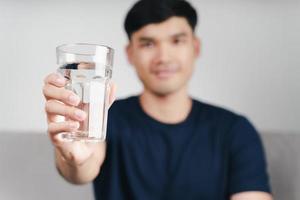 knappe aziatische man die een glas water drinkt op de bank in de woonkamer foto