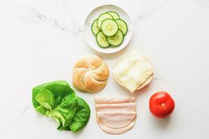 voedsel producten en ingrediënten voor maken Sandwich. ham, kaas, hamburger broodje, sla, komkommer en tomaat net zo recept flatlay Aan marmeren keuken tafel foto