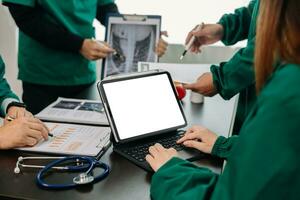 medisch team hebben een vergadering met artsen in wit laboratorium jassen en chirurgisch scrubs gezeten Bij een tafel bespreken een patiënten werken online gebruik makend van computers in de medisch foto