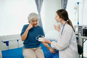 dokter in wit pak nemen aantekeningen terwijl bespreken en Aziatisch ouderen, vrouw geduldig wie aan het liegen Aan bed met ontvangen zoutoplossing oplossing in ziekenhuis of kliniek. foto