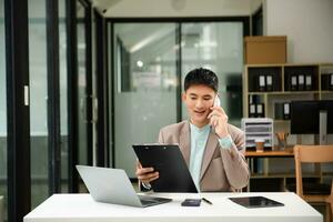 Aziatisch wman pratend Aan de telefoon met een klant jong positief vrouw accountant gebruik makend van smartphone pratend naar team Bij haar bureau ontspannende met mobiel toepassing foto