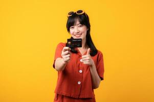 portret jong mooi Aziatisch vrouw gelukkig glimlach gekleed in oranje kleren tonen credit kaart en duim vinger omhoog gebaar geïsoleerd Aan geel achtergrond. betalen en aankoop boodschappen doen betaling concept. foto