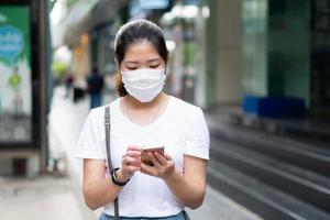 jonge aziatische vrouw die gezichtsmasker draagt die smartphone gebruikt en in de stad loopt tijdens covid-19 of coronavirusuitbraak. sociale afstand en nieuw normaal levensstijlconcept lifestyle
