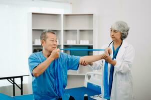 Aziatisch fysiotherapeut helpen ouderen Mens geduldig uitrekken arm gedurende oefening correct met halter in hand- gedurende opleiding hand- in bed in kliniek foto