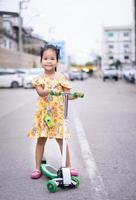 schattig klein Aziatisch meisje in gele jurk die een scooter op straat speelt foto