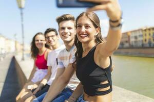 Italië, pisa, groep van vier gelukkig vrienden zittend Aan een muur langs arno rivier- nemen een selfie foto