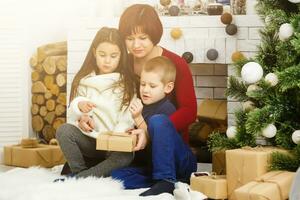 familie op kerstavond bij open haard. moeder en kleine kinderen openen kerstcadeautjes. kinderen met geschenkdozen. woonkamer met traditionele open haard en versierde boom. gezellige winteravond thuis. foto