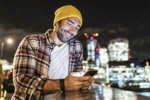 uk, Londen, glimlachen Mens leunend Aan een traliewerk en op zoek Bij zijn telefoon met stad lichten in achtergrond foto