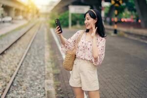 Aziatisch jong vrouw reiziger met het weven mand gebruik makend van een mobiel telefoon naast spoorweg trein station in Bangkok. reis reis levensstijl, wereld reizen ontdekkingsreiziger of Azië zomer toerisme concept. foto