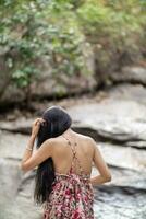 jong Dames toerist in wijnoogst jurk staat alleen Aan rots door waterval in de Woud hopen naar koel naar beneden voor zwemmen. de terug van jong Aziatisch Dames toerist wie geniet spelen in waterval alleen. foto