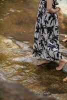 jong Dames toerist in wijnoogst jurk staat alleen Aan rots door waterval in de Woud hopen naar koel naar beneden voor zwemmen. de terug van jong Aziatisch Dames toerist wie geniet spelen in waterval alleen. foto