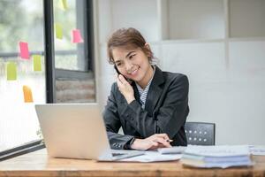 Aziatisch zakenvrouw pratend Aan mobiel telefoon werken Aan laptop in modern kantoor. gelukkig bedrijf vrouw pratend Aan mobiel telefoon terwijl analyseren wekelijks schema in laptop foto