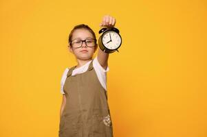 een zwart alarm klok in meisjes hand, tonen tijd Aan de bellen, geïsoleerd oranje achtergrond. terug naar school. tijd beheer foto