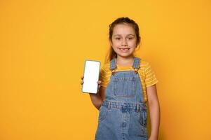 aanbiddelijk meisje gekleed in geel t-shirt en denim overall, tonen een slim mobiel telefoon met wit leeg blanco scherm foto