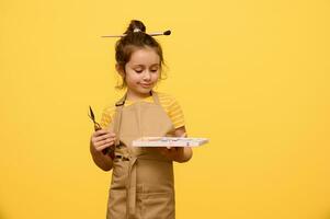 portret van een Kaukasisch schattig weinig kind meisje, schilder artiest met schilderij hulpmiddelen, geïsoleerd Aan geel studio achtergrond foto