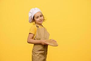 schattig weinig chef banketbakker, koken, bakker, pizzaiolo, aanbiddelijk kind meisje houdt een houten bord, glimlacht Bij camera foto