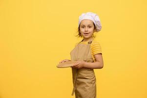 aanbiddelijk kind schoolmeisje gekleed net zo chef banketbakker, houdt een houten bord, glimlacht op zoek Bij camera, geïsoleerd Aan geel foto