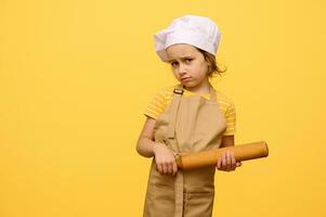 weinig peuter- meisje met rollend pin, gekleed net zo chef gebakje, op zoek ongelukkig, geïsoleerd over- geel studio achtergrond foto