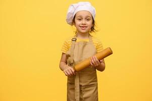 gelukkig weinig peuter- meisje met rollend pin, gekleed net zo chef gebakje, glimlachen op zoek Bij camera, geel achtergrond foto