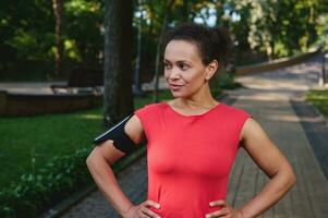 charmant gespierd vrouw met gezond esthetisch lichaam en gloeiend huid, atleet in rood t-shirt en smartphone houder poseren met armen Aan taille op zoek terzijde staand Aan de stad park loopband foto