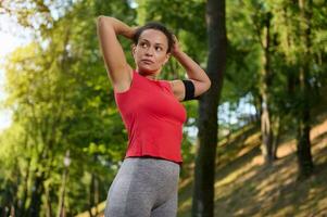 zelfverzekerd mooi fit vrouw in sportkleding met een smartphone houder koppelverkoop een paardenstaart, staand in een stad park Aan een zonnig warm zomer dag en krijgen klaar voor een rennen in de park foto