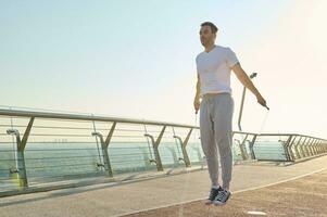 vol lengte portret van een gespierd bouwen Kaukasisch Mens, fit atleet aan het doen jumping opdrachten, cardio opleiding met een overslaan touw Aan de glas stad brug vroeg in de ochtend- Aan een zomer dag foto