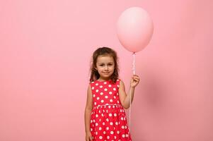 portret van een mooi mooi prachtig aanbiddelijk 4 jaren verjaardag meisje, kind in jurk met polka stippen patroon, Holding een roze ballon, geïsoleerd Aan roze achtergrond met kopiëren ruimte voor reclame. foto