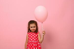 portret van mooi mooi prachtig aanbiddelijk 4 jaren verjaardag meisje, kind in jurk met polka stippen patroon, Holding een roze ballon, geïsoleerd over- roze achtergrond met kopiëren ruimte voor reclame. foto