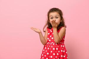 prachtig kind meisje in roze jurk met polka dots stuurt een lucht kus, op zoek Bij de camera, poseren Aan een roze achtergrond met kopiëren ruimte. portret van een mooi kind met positief emoties voor reclame foto