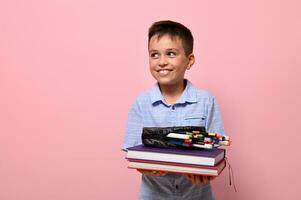 een charmant jongen, een leerling Bij school, houdt boeken in voorkant van hem en potlood geval met pennen, viltstift pennen en markeringen vallend uit van hem. school- jongen schattig glimlacht op zoek naar de kant Aan roze achtergrond foto