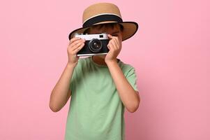 schooljongen in groen t-shirt en zomer hoed nemen foto gebruik makend van een oud wijnoogst retro camera, geïsoleerd over- roze achtergrond met kopiëren ruimte. toerisme concepten