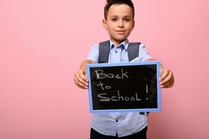 aanbiddelijk schooljongen met rugzak houdt schoolbord met krijt belettering ,rug naar school. geïsoleerd over- roze achtergrond met ruimte voor tekst foto